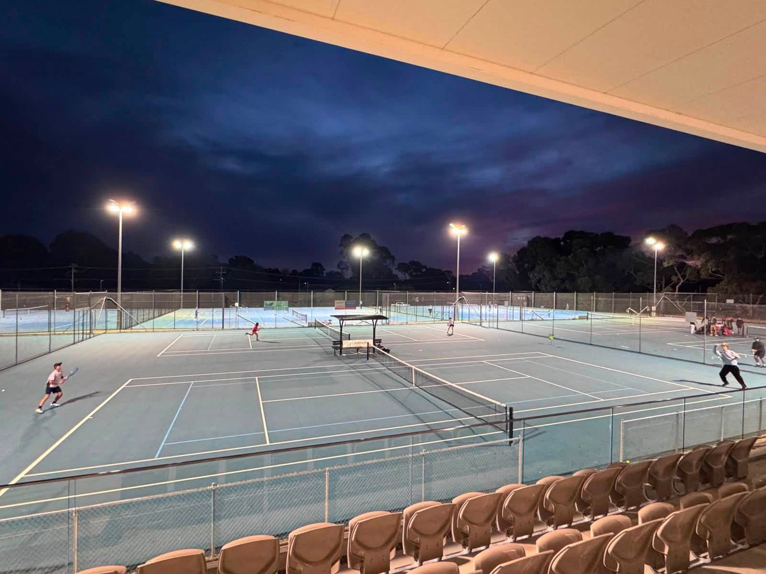 Four floodlit hard courts at Tea Tree Gully at dusk, with players rallying under the lights and the last of the sunset behind the gum trees.