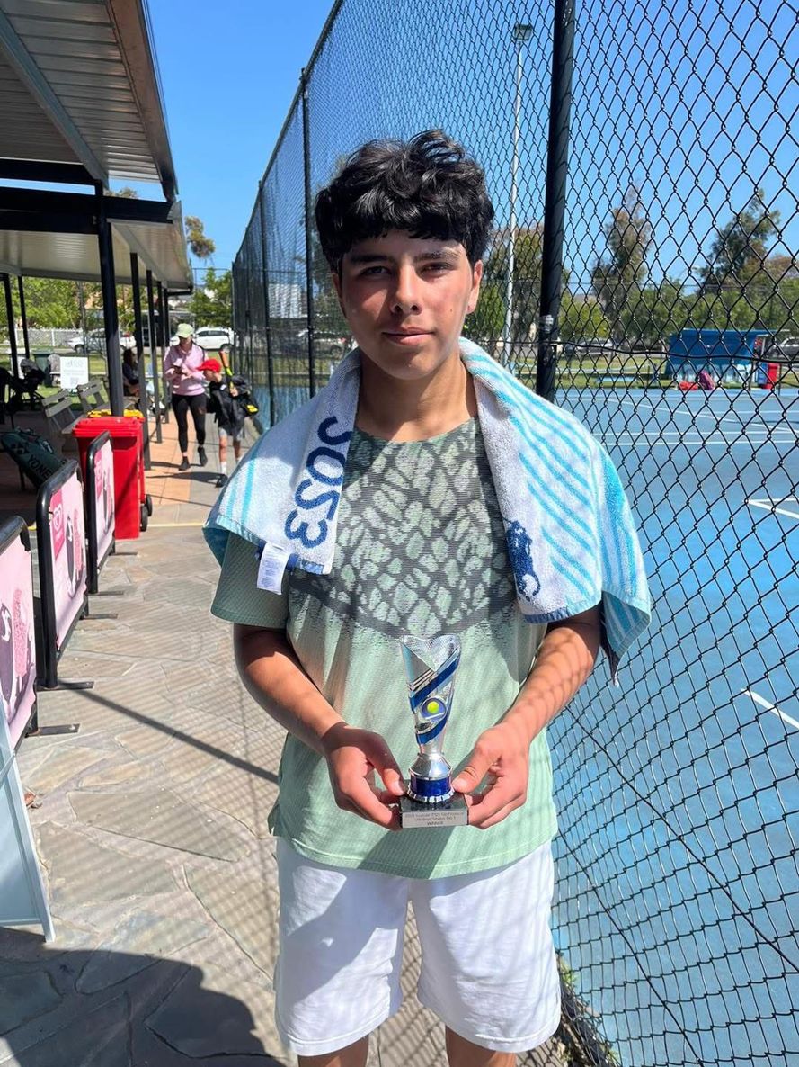 A junior player holding a trophy behind the court fence at an outdoor tournament venue