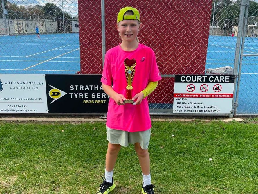 A junior player holding a trophy on the grass beside the courts