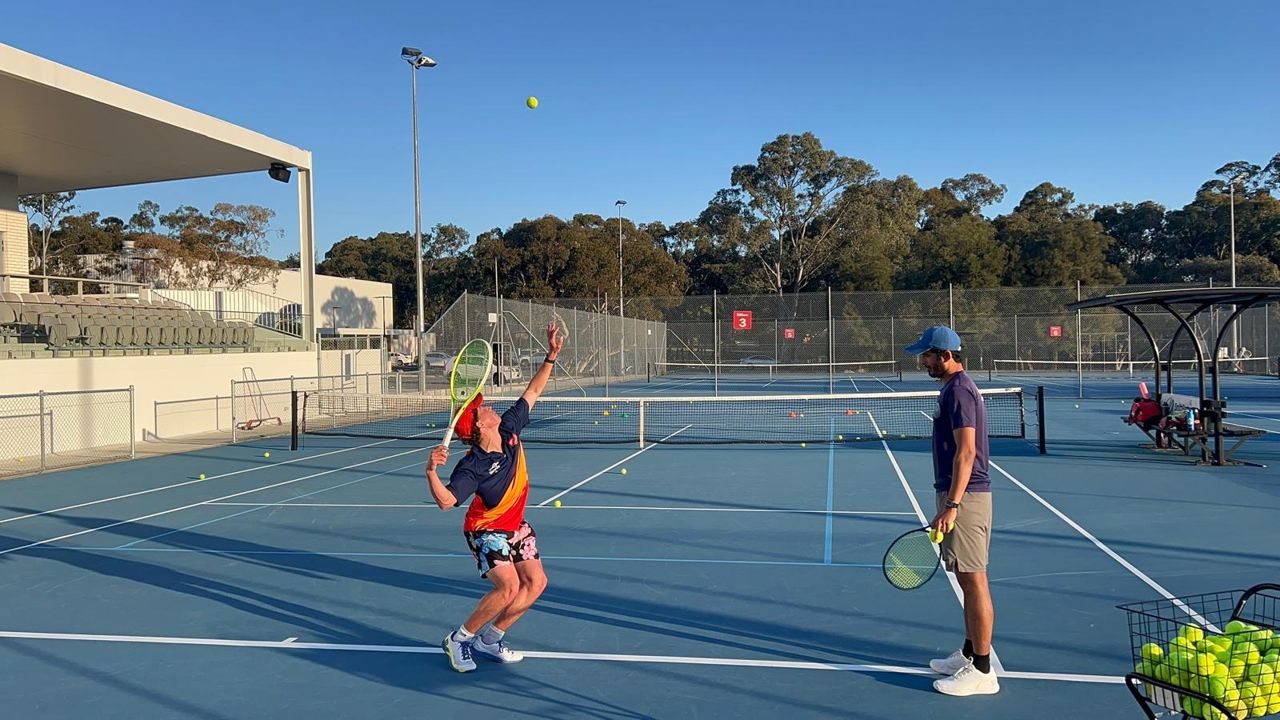 A player practising serves under a coach's watch on an outdoor hard court, with a ball basket courtside