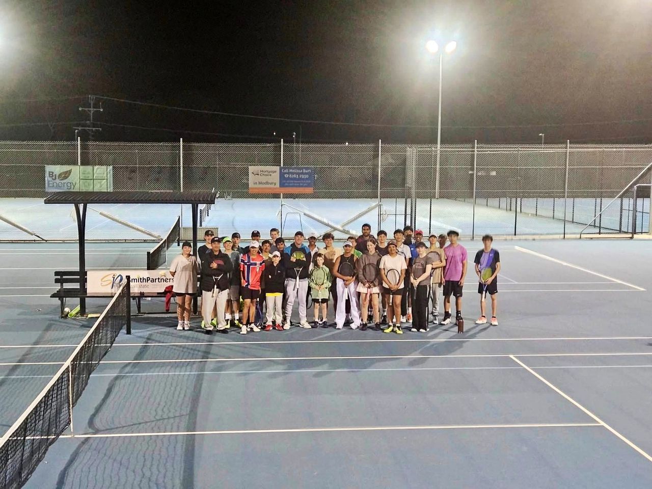 A camp group and coaches lined up along the net under floodlights
