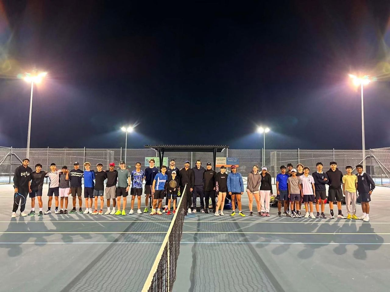 Squad players lined up along the net on floodlit courts at the end of an evening session